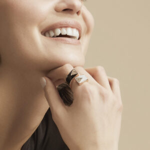 anello-cristalllo-amplecti Smiling woman with designer Amplecti gold ring with inset diamonds on neutral background