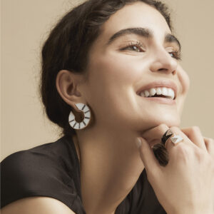 gioielli-cristalllo-anello-orecchini Happy young woman with Cavea temple vesta circular design and modern gold Amplecti ring, posing for a jewelry photo on a neutral background